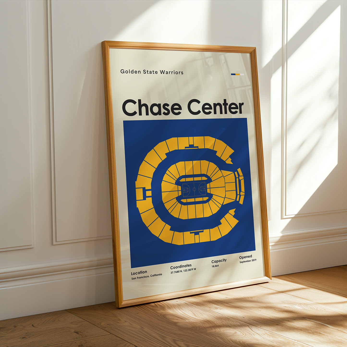 Framed poster of the Golden State Warriors Chase Center on a wooden floor with sunlight streaming in.