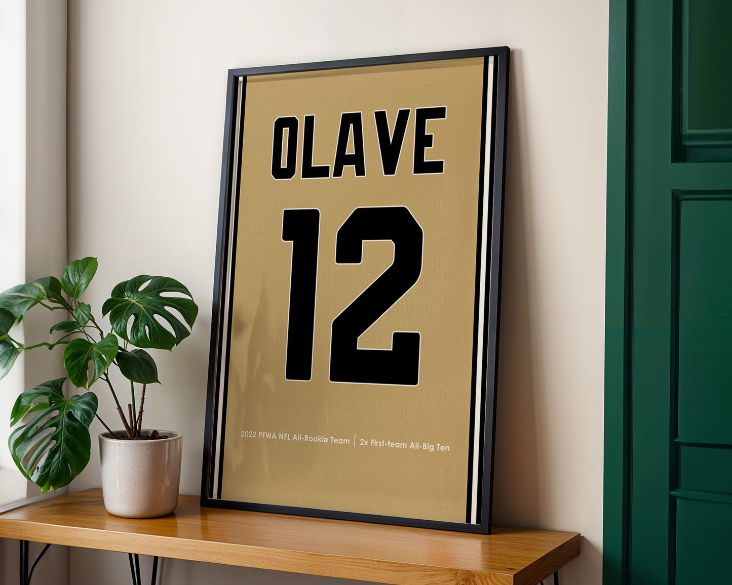 Framed poster with 'OLAVE 12' on a wooden shelf next to a plant.