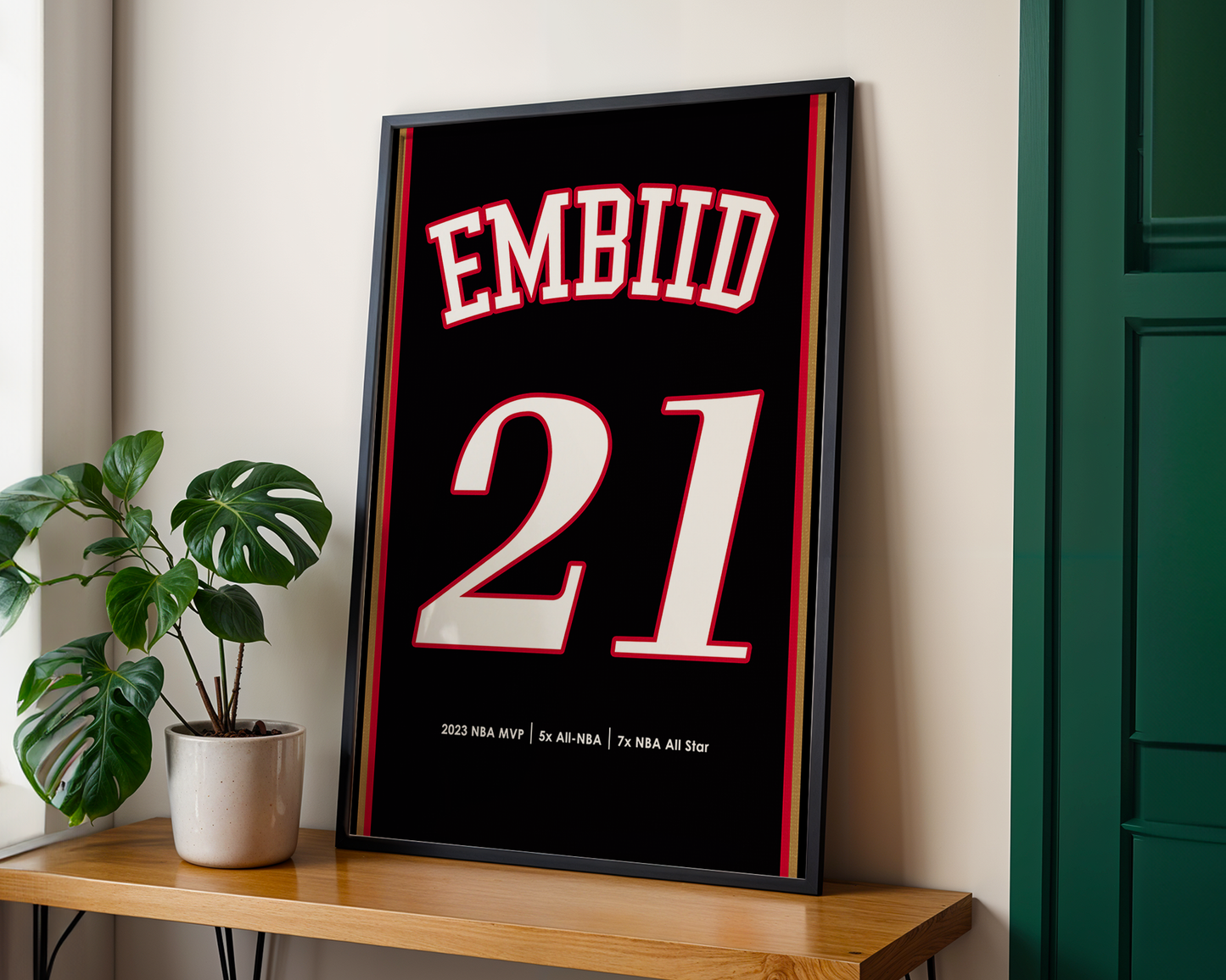 Framed sports jersey with 'EMBIID 21' on a wooden shelf next to a plant.