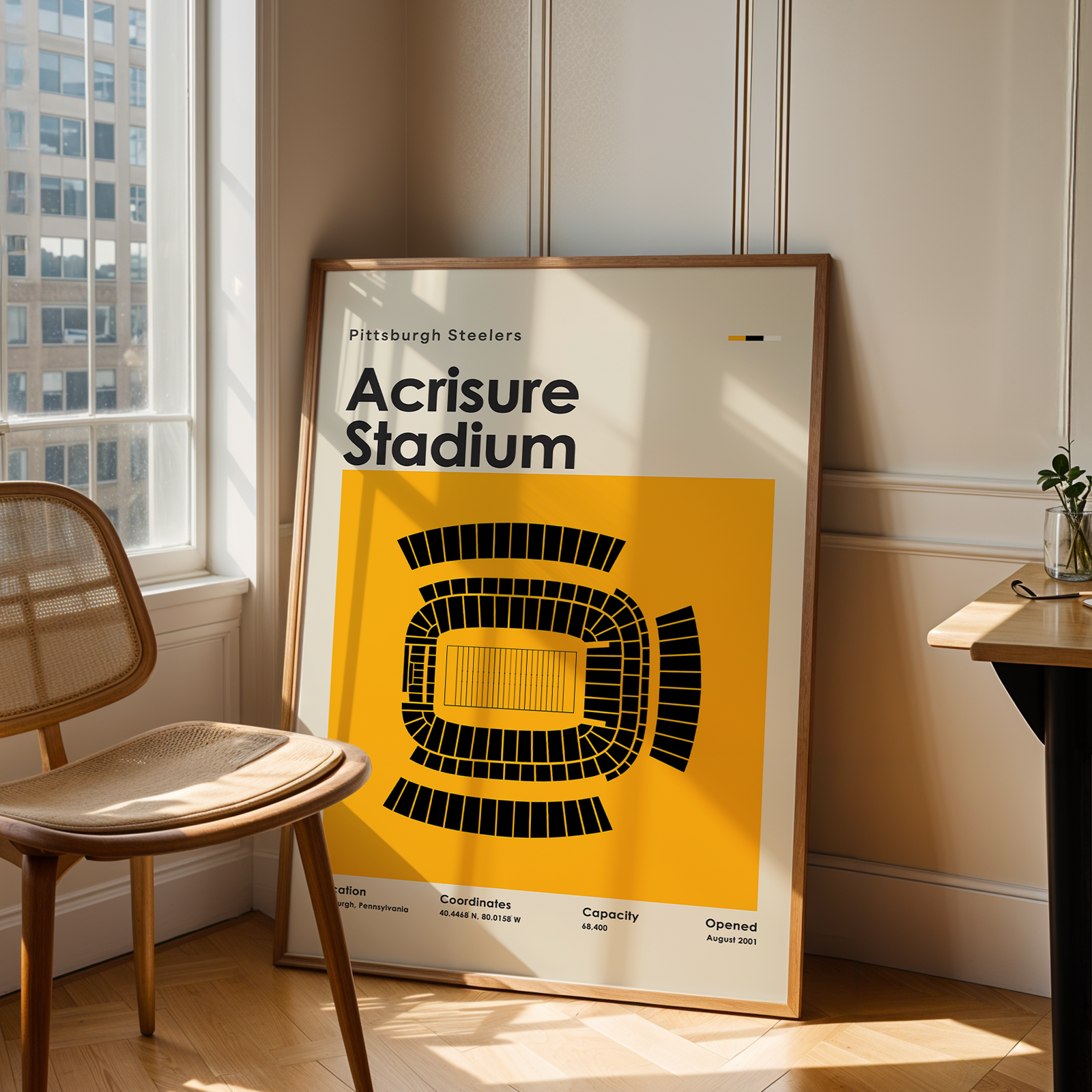 Framed poster of Acrisure Stadium with Pittsburgh Steelers logo in a room with a window and chair.
