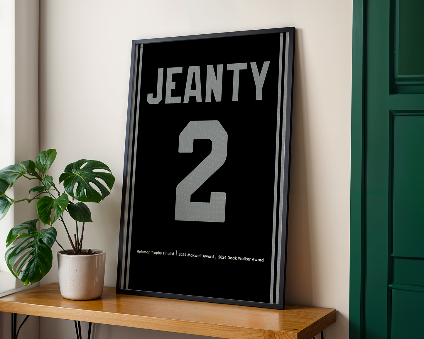 Framed poster with 'JEANTY' and number '2' on a wall next to a plant and wooden shelf.