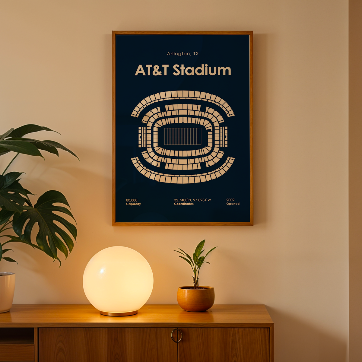 A framed AT&T Stadium print hangs on a beige wall above a wooden console table. The poster is dark blue with a cream stadium map and text, surrounded by a warm glowing spherical lamp and potted plants.