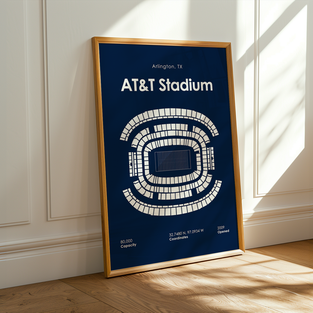 A framed AT&T Stadium print leaning against a white paneled wall on a light wood floor, with warm sunlight casting shadows across the wall and floor.