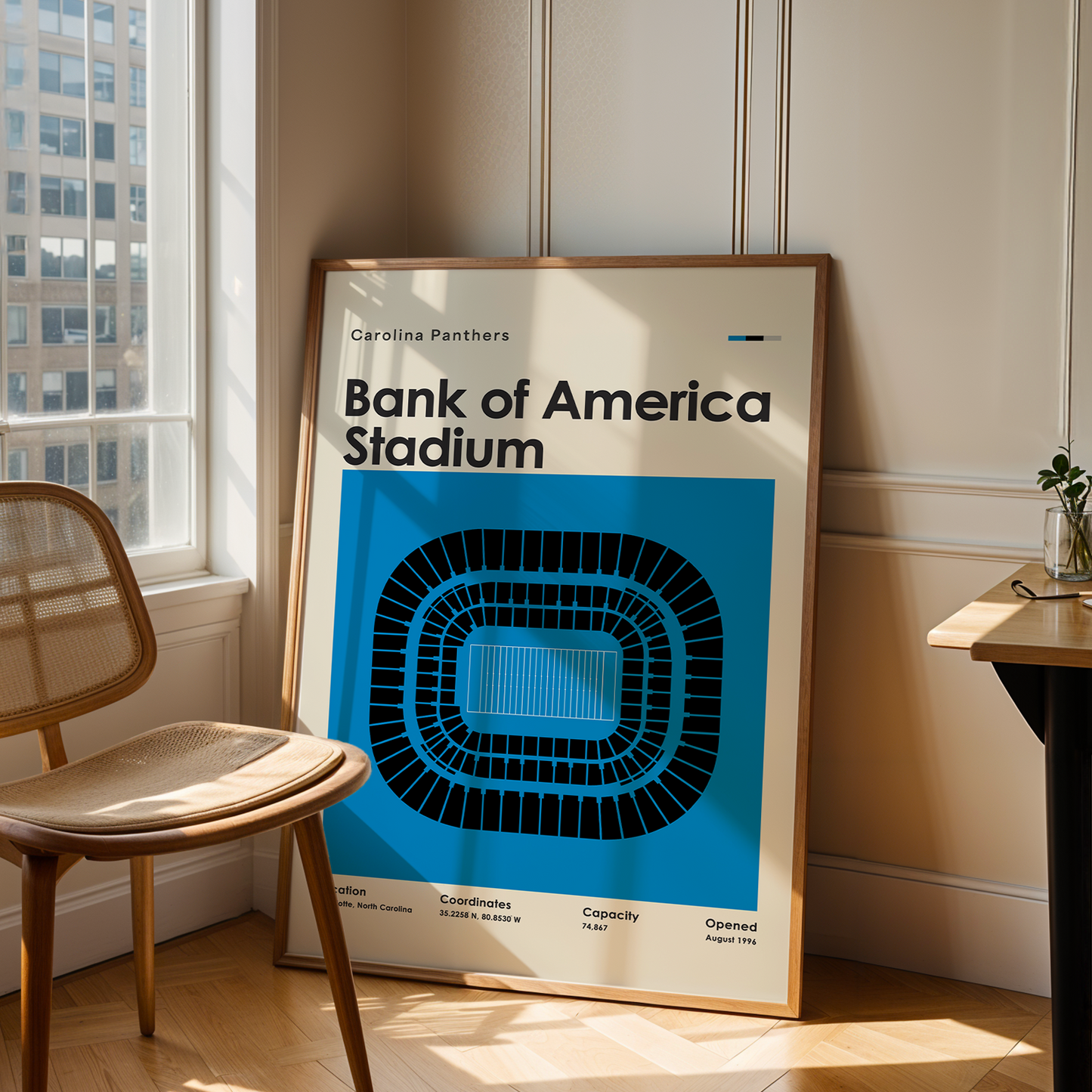 Framed poster of Bank of America Stadium in a room with a chair and table.