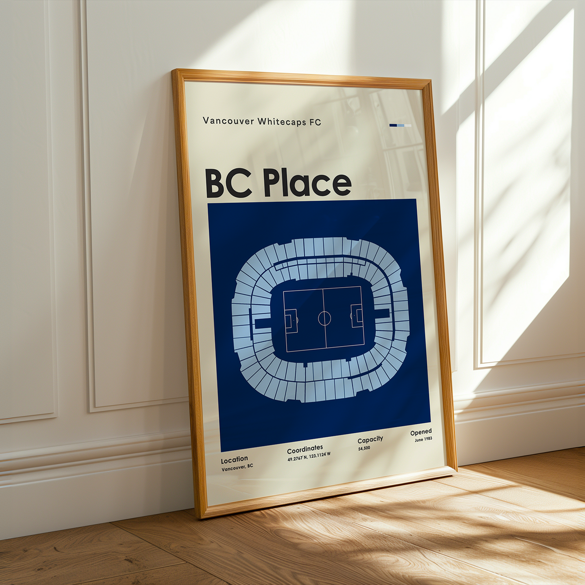 A framed Vancouver Whitecaps FC BC Place sports poster leaning against a white paneled wall on a light wood floor, with warm sunlight streaming in from the right.