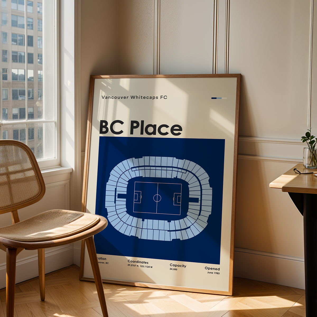 A framed BC Place soccer stadium graphic print leaning against a wall in a sunlit room next to a wooden chair and desk.
