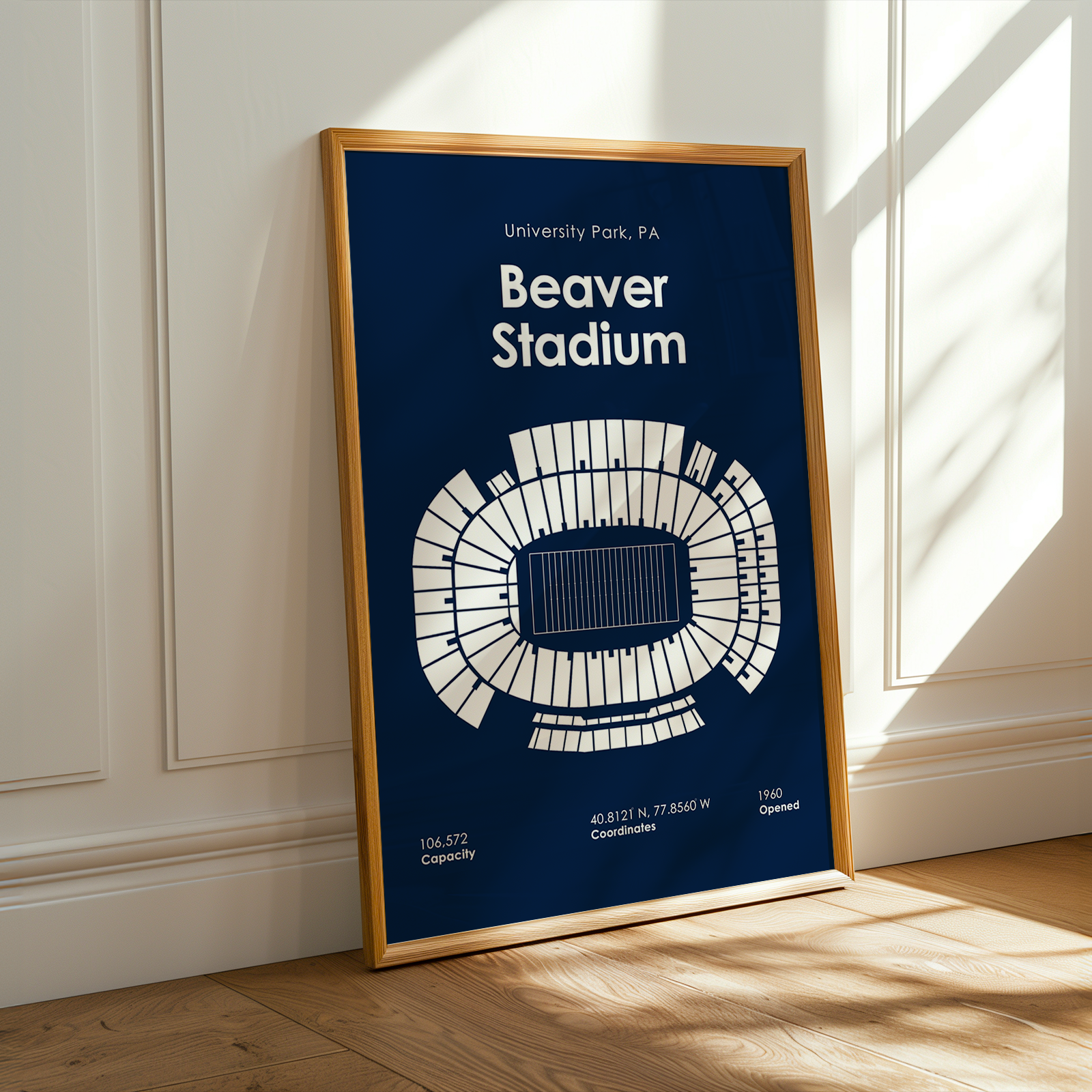 A framed poster of University Park Stadium in Beaver Stadium, PA, leaning against a white paneled wall on a light wood floor with warm sunlight streaming in from the side.