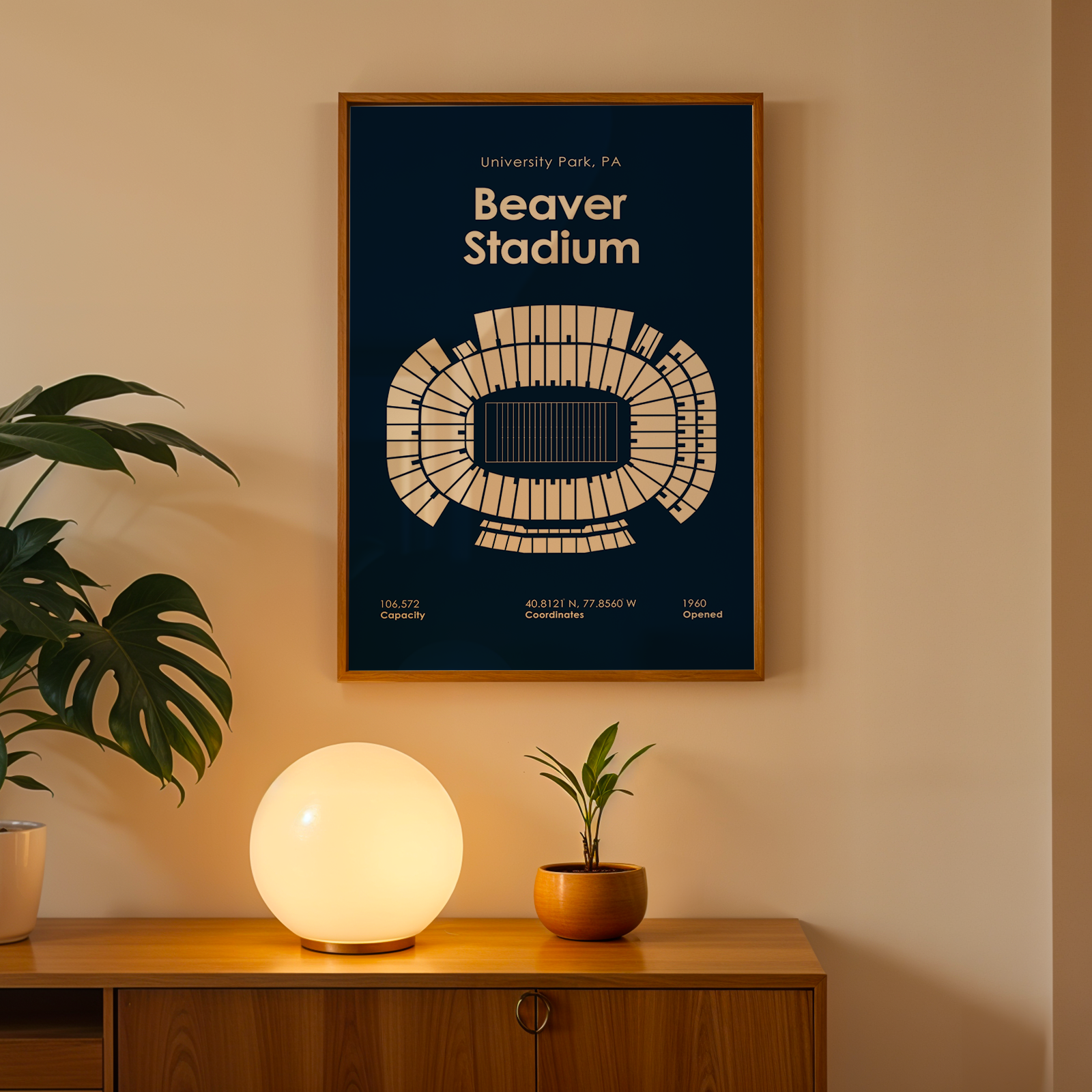 A framed poster of Beaver Stadium in University Park, PA, hanging on a beige wall above a wooden console table with a glowing globe lamp and potted plants.
