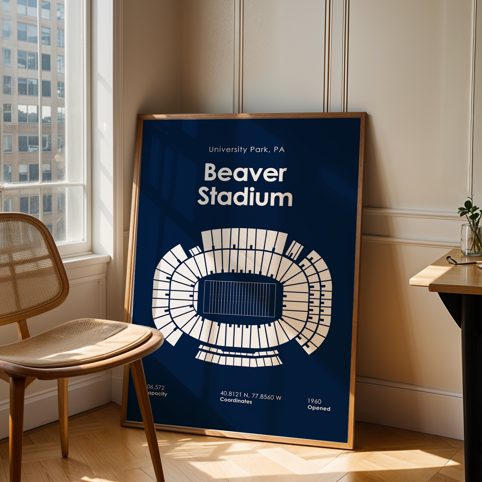 A framed poster of Beaver Stadium in University Park, PA, leaning against a wall in a modern room with a wooden chair and desk.
