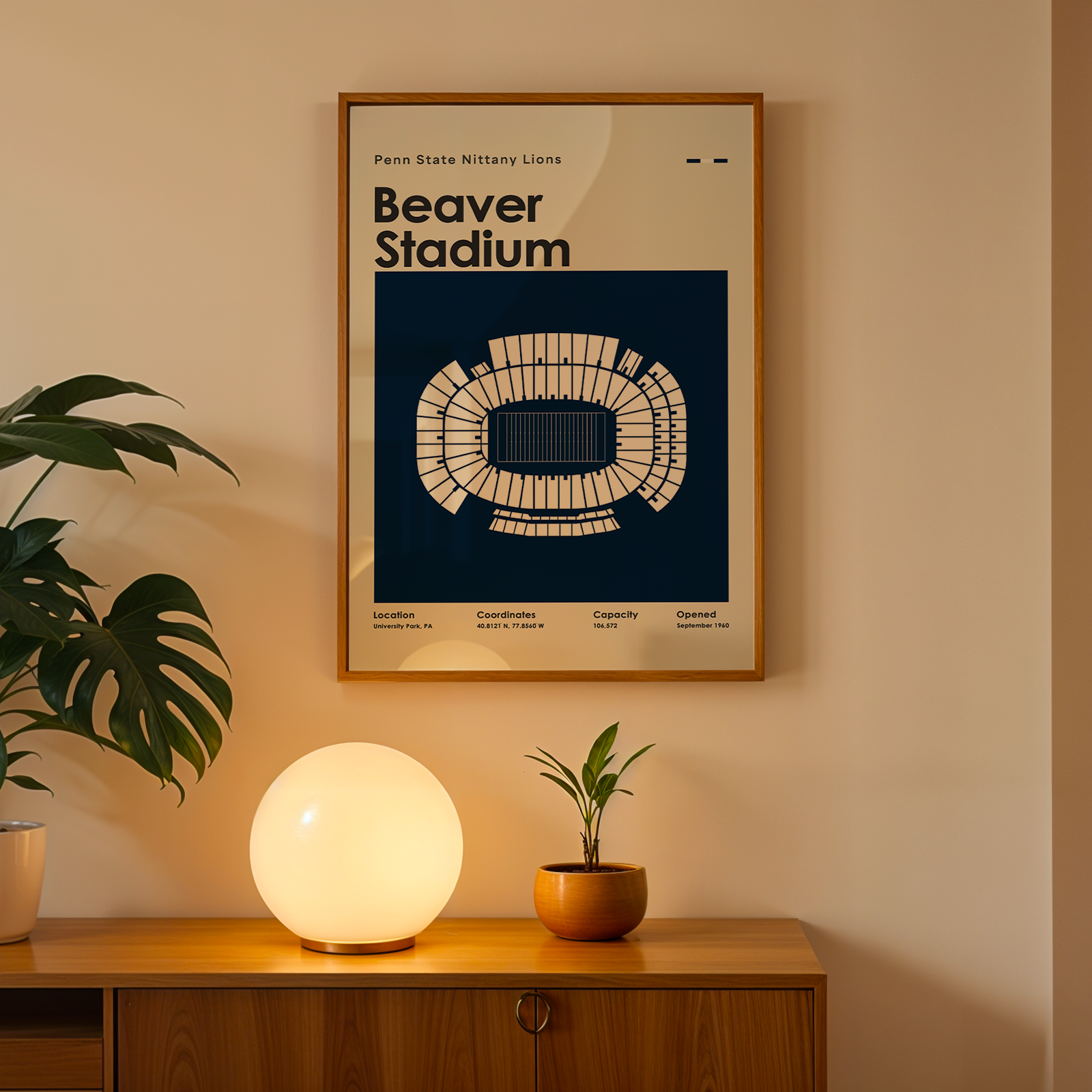 A framed poster of Beaver Stadium in Mountaineer Blue, featuring a white stadium diagram on a navy blue background, hanging on a beige wall above a wooden console table with a glowing globe lamp and potted plants.