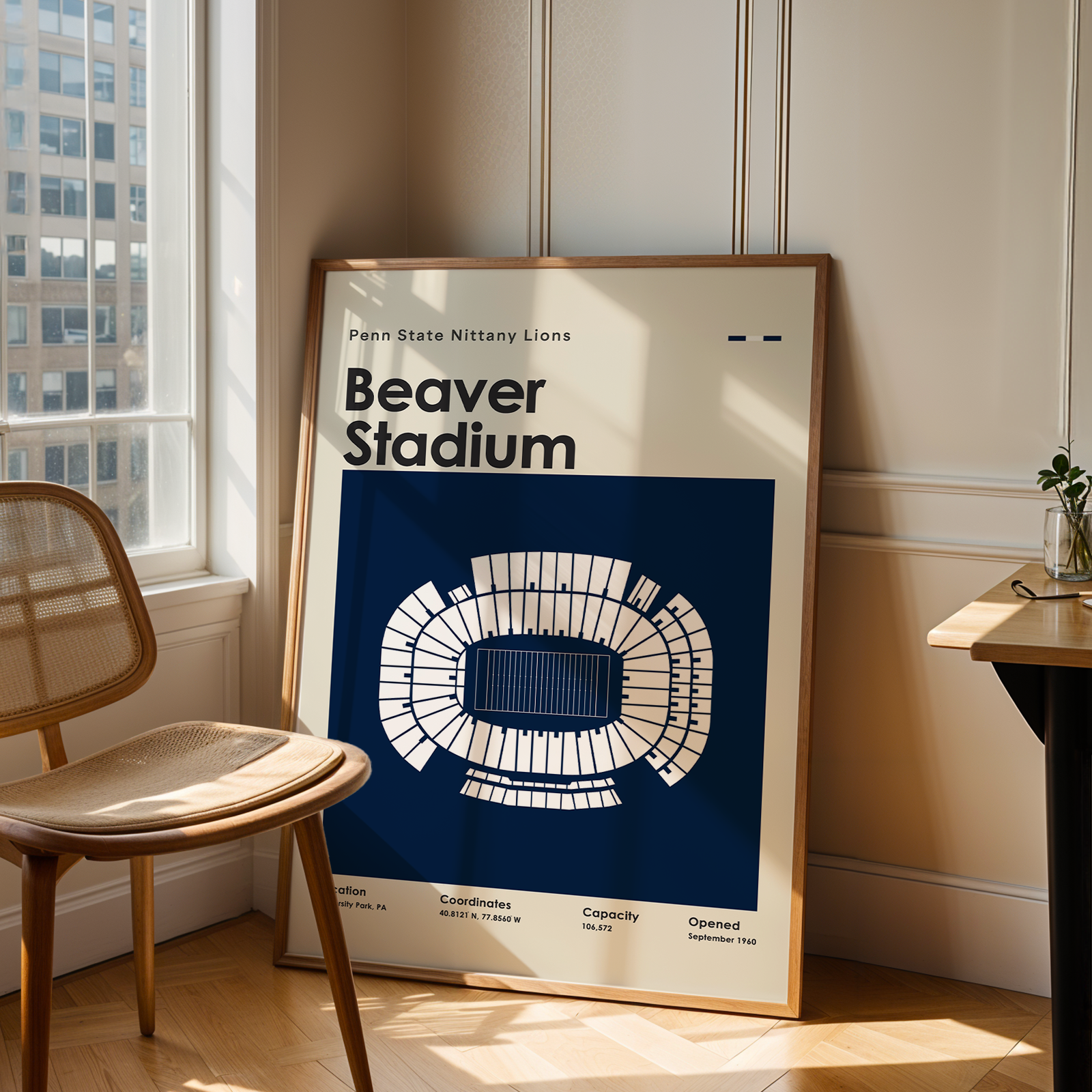 A framed poster of Beaver Stadium in Pittsburgh, featuring a blue and cream stadium diagram, leaning against a wall in a sunlit room next to a wooden chair and desk.