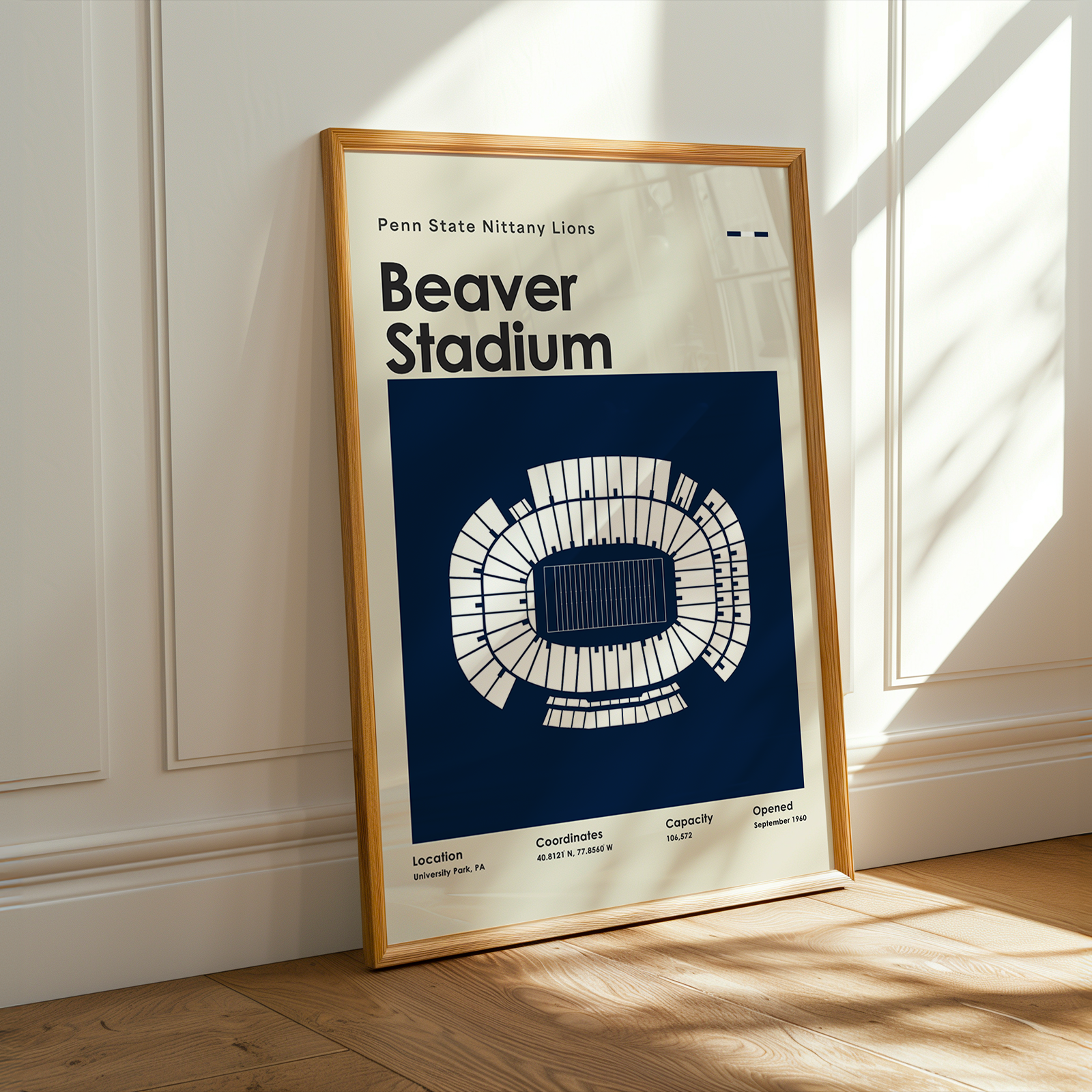 A framed poster of Penn State Nittany Lions Beaver Stadium leaning against a white paneled wall on a light wood floor, with warm sunlight casting shadows across the scene.