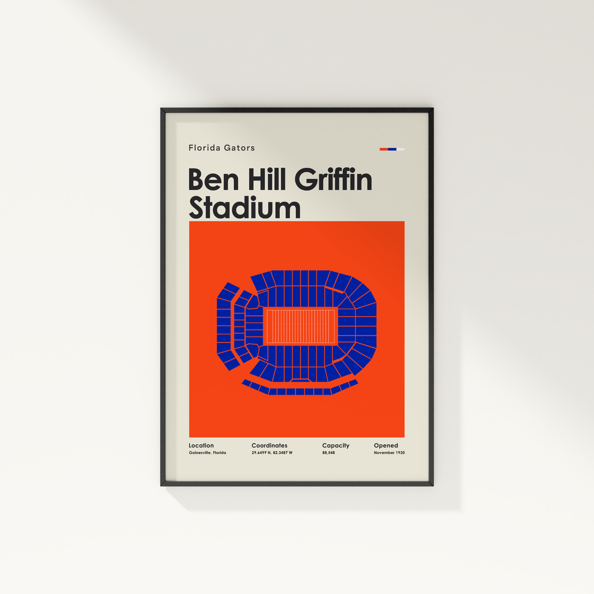 A framed poster of Ben Hill Griffin Stadium in Florida Gators branding, featuring a minimalist design with an orange background, blue stadium layout, and black text, displayed on a clean white wall.