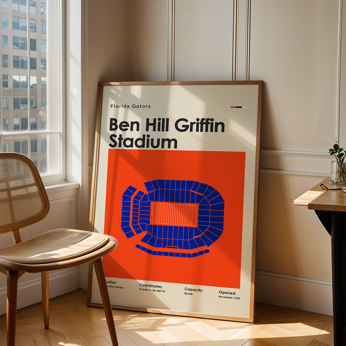 A framed poster of Ben Hill Griffin Stadium featuring a bold orange and blue graphic, leaning against a wall in a sunlit room next to a wooden chair and desk.