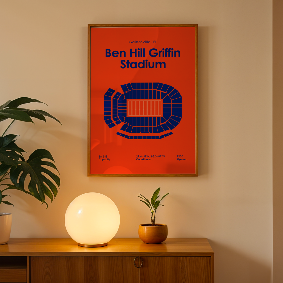 A framed poster of Ben Hill Griffin Stadium in orange and blue hanging on a beige wall above a wooden console table with a glowing globe lamp and potted plants.