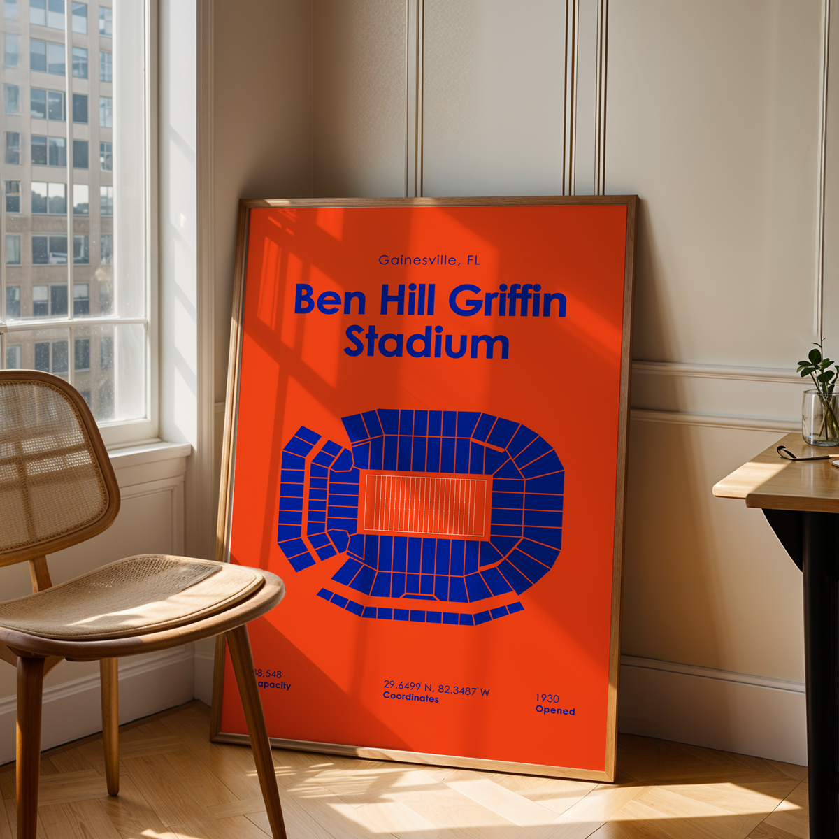 A framed orange and blue poster of Ben Hill Griffin Stadium in Gainesville, Florida, leaning against a wall in a sunlit room next to a wooden chair and desk.