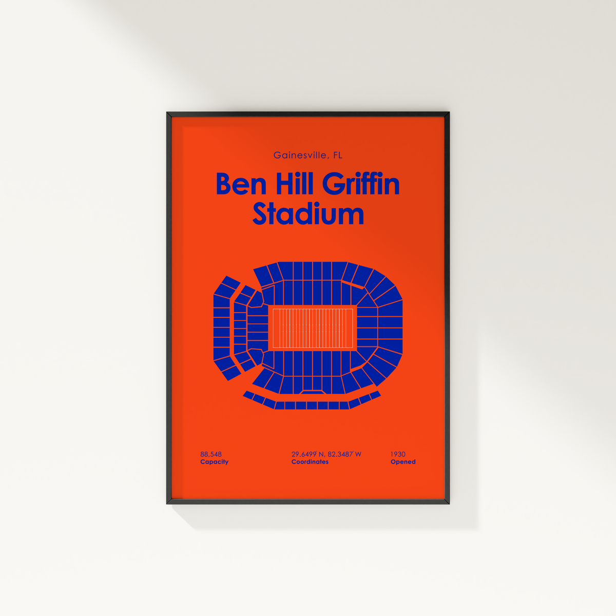 A framed sports poster of Ben Hill Griffin Stadium in Gainesville, FL, featuring a bold orange background, blue stadium diagram, and blue text, shown hanging on a white wall.