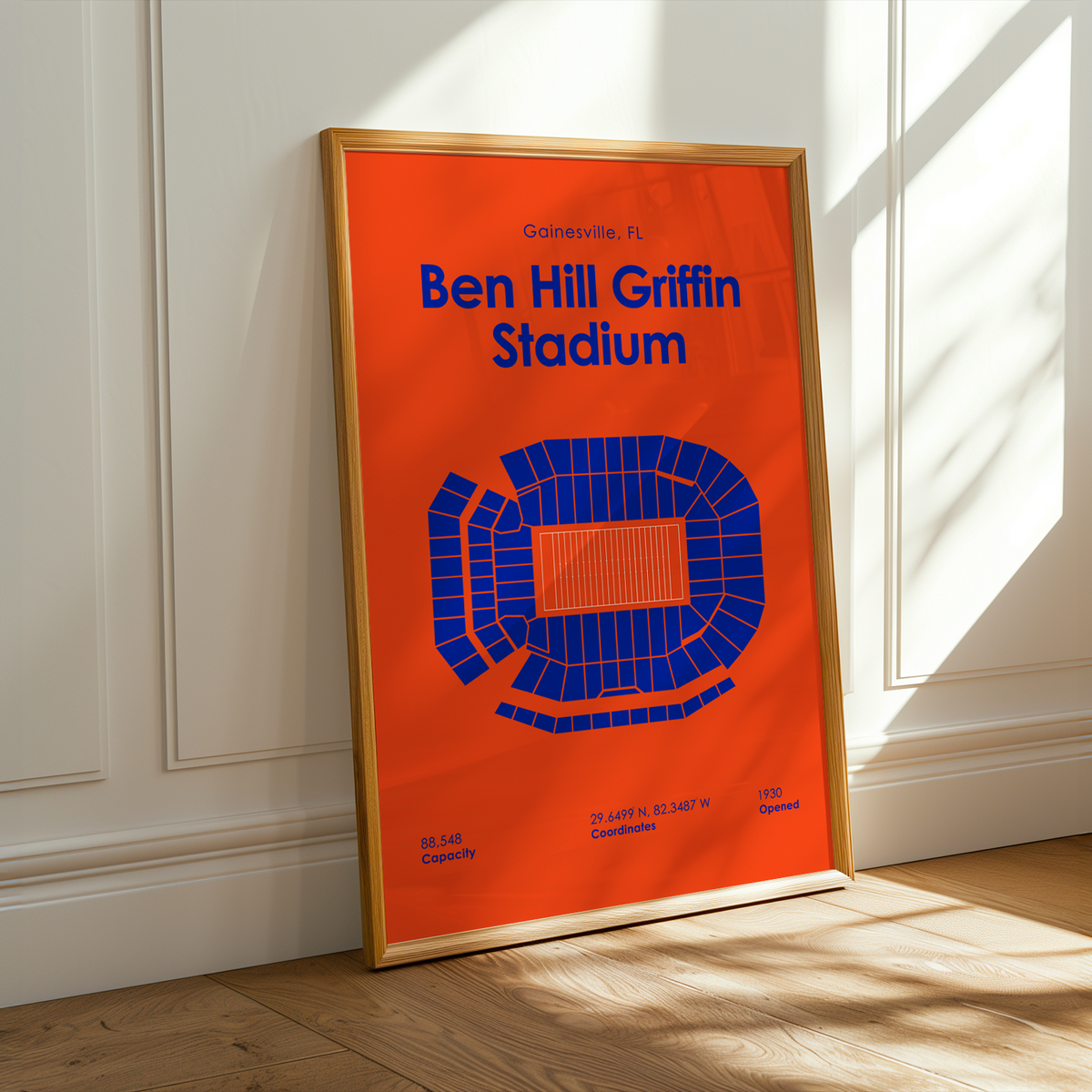 A framed sports poster of Ben Hill Griffin Stadium in Gambelle, FL, leaning against a white paneled wall on a light wood floor with warm sunlight streaming in.