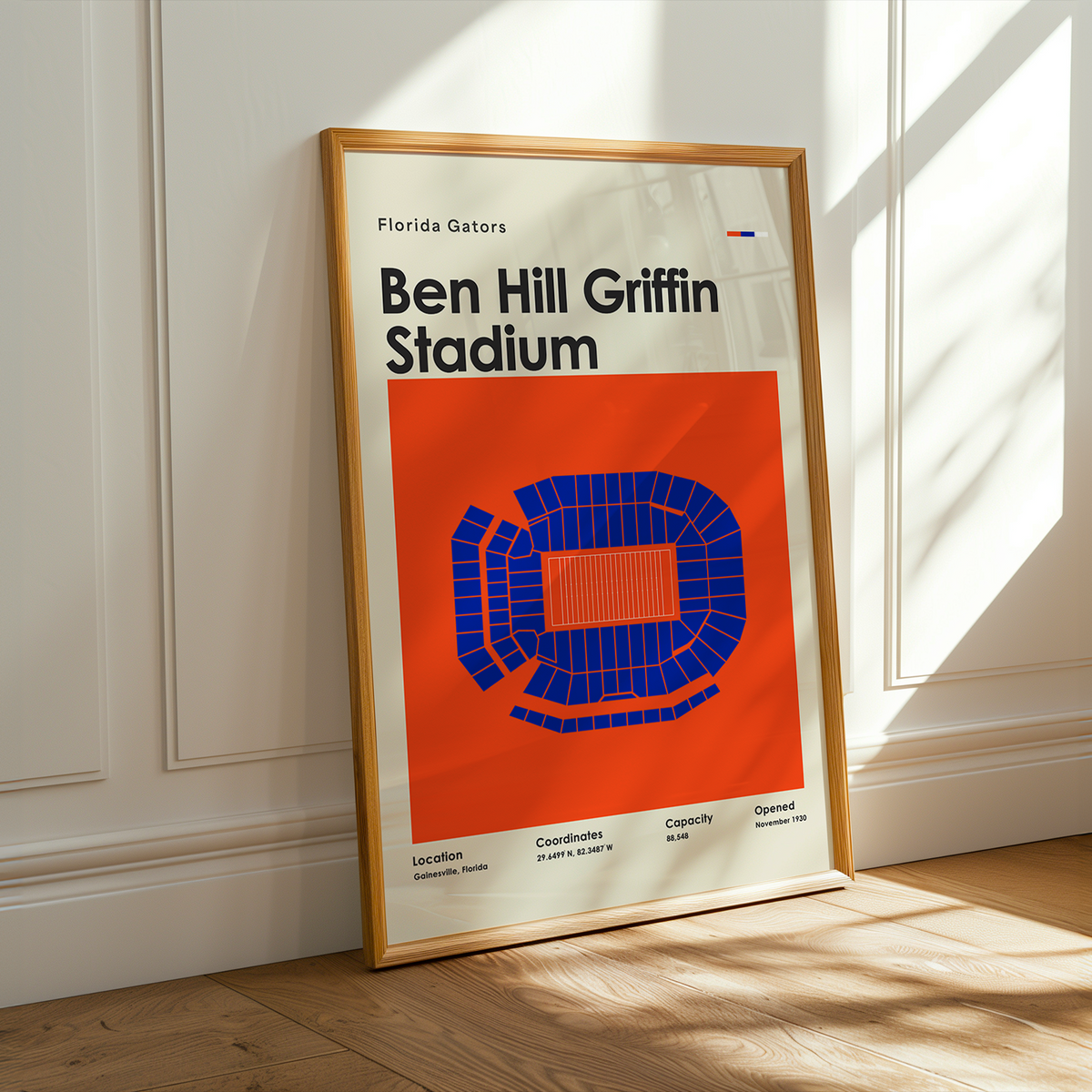 A framed poster of Ben Hill Griffin Stadium from the Florida Gators, leaning against a white paneled wall on a wooden floor with warm sunlight streaming in.