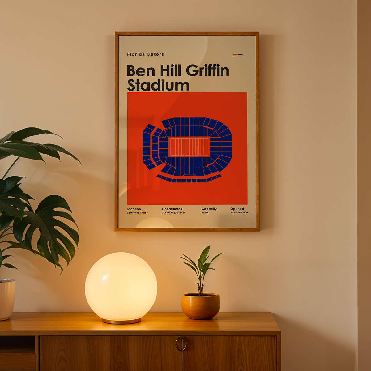 A framed Ben Hill Griffin Stadium poster hangs on a beige wall above a wooden console table. The poster features a bold red-orange background with a blue stadium layout and black text.