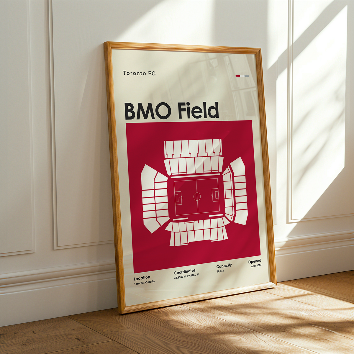 A framed BMO Field poster leaning against a white paneled wall on a light wood floor, with warm sunlight casting shadows across the scene.