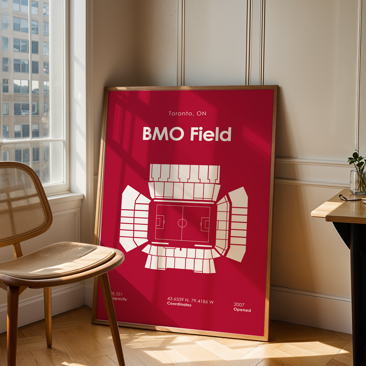 A framed minimalist stadium map poster of BMO Field in Toronto, leaning against a beige paneled wall in a sunlit room next to a wooden chair and table.
