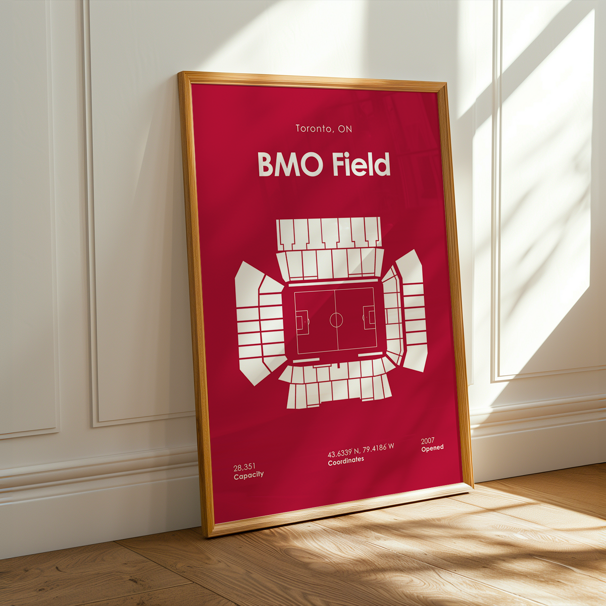 A framed minimalist sports poster of BMO Field in Toronto, leaning against a white paneled wall on a light wood floor with warm sunlight casting shadows.