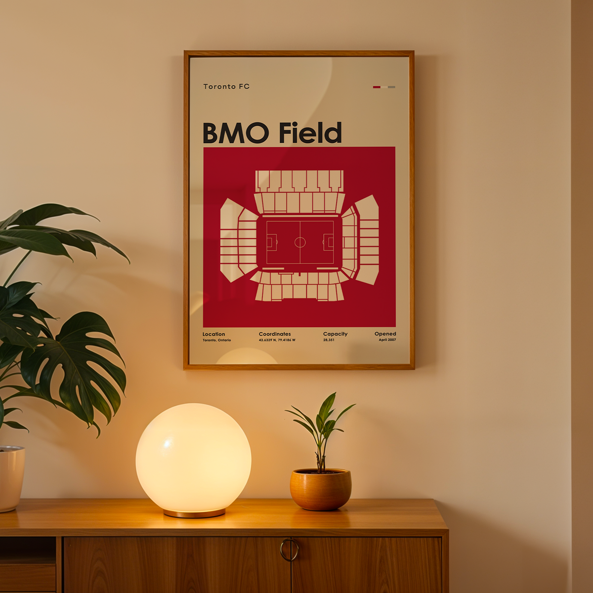 A framed BMO Field poster hangs on a beige wall above a wooden console table. The poster features a bold red and cream stadium map design with the text 'BMO Field' and 'Toronto FC'.