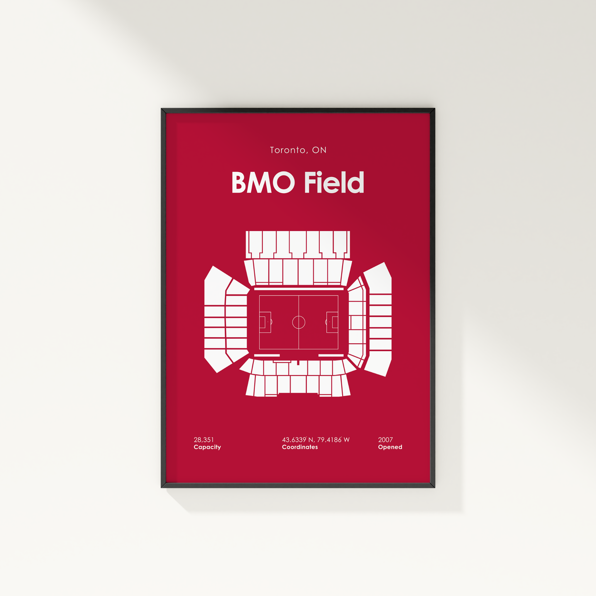 A framed minimalist sports poster of BMO Field in Toronto, featuring a white stadium floor plan and text on a deep red background, shown hanging on a light gray wall.