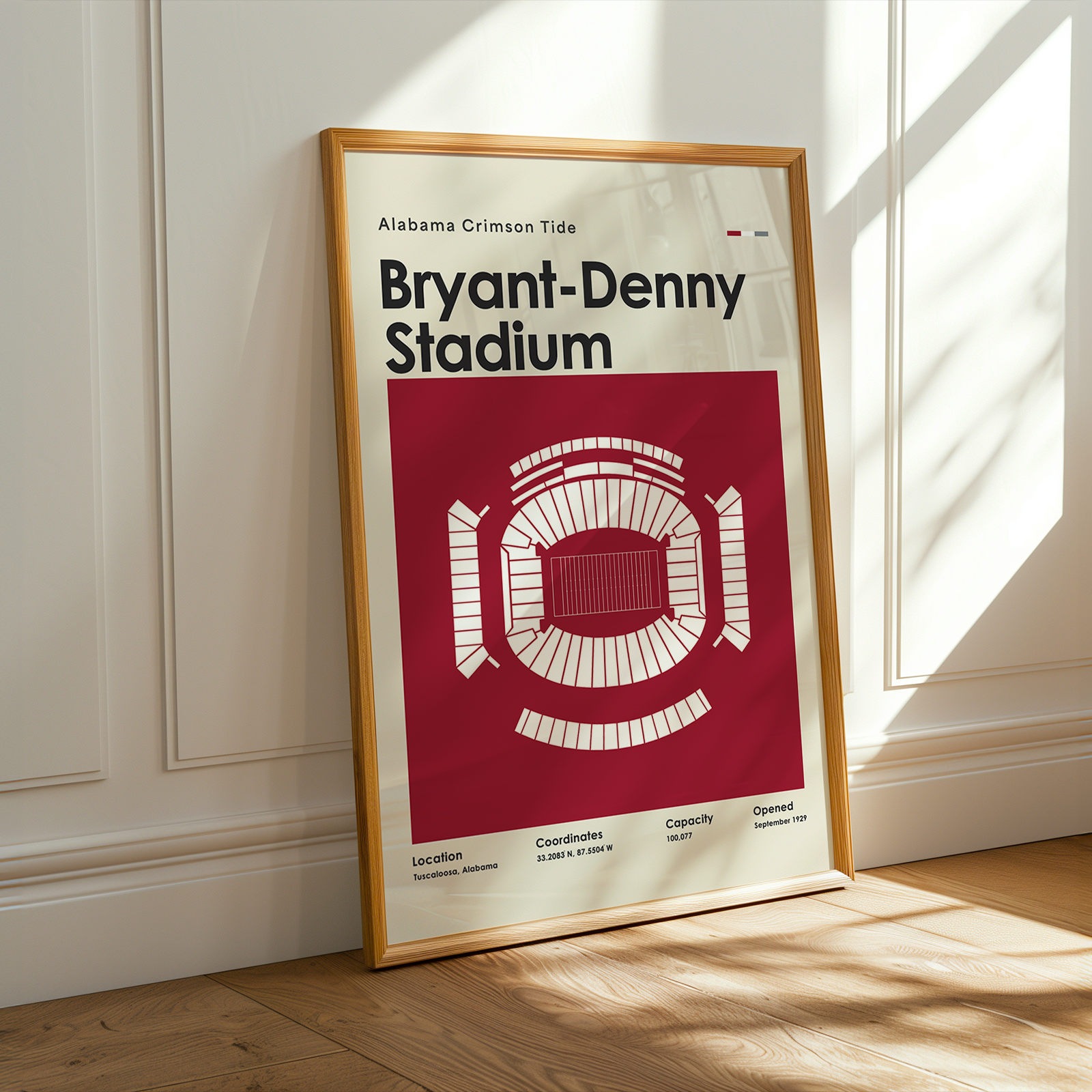 A framed poster of Bryant-Denny Stadium, Alabama Crimson Tide, leaning against a white paneled wall on a wooden floor with warm sunlight and window shadows.