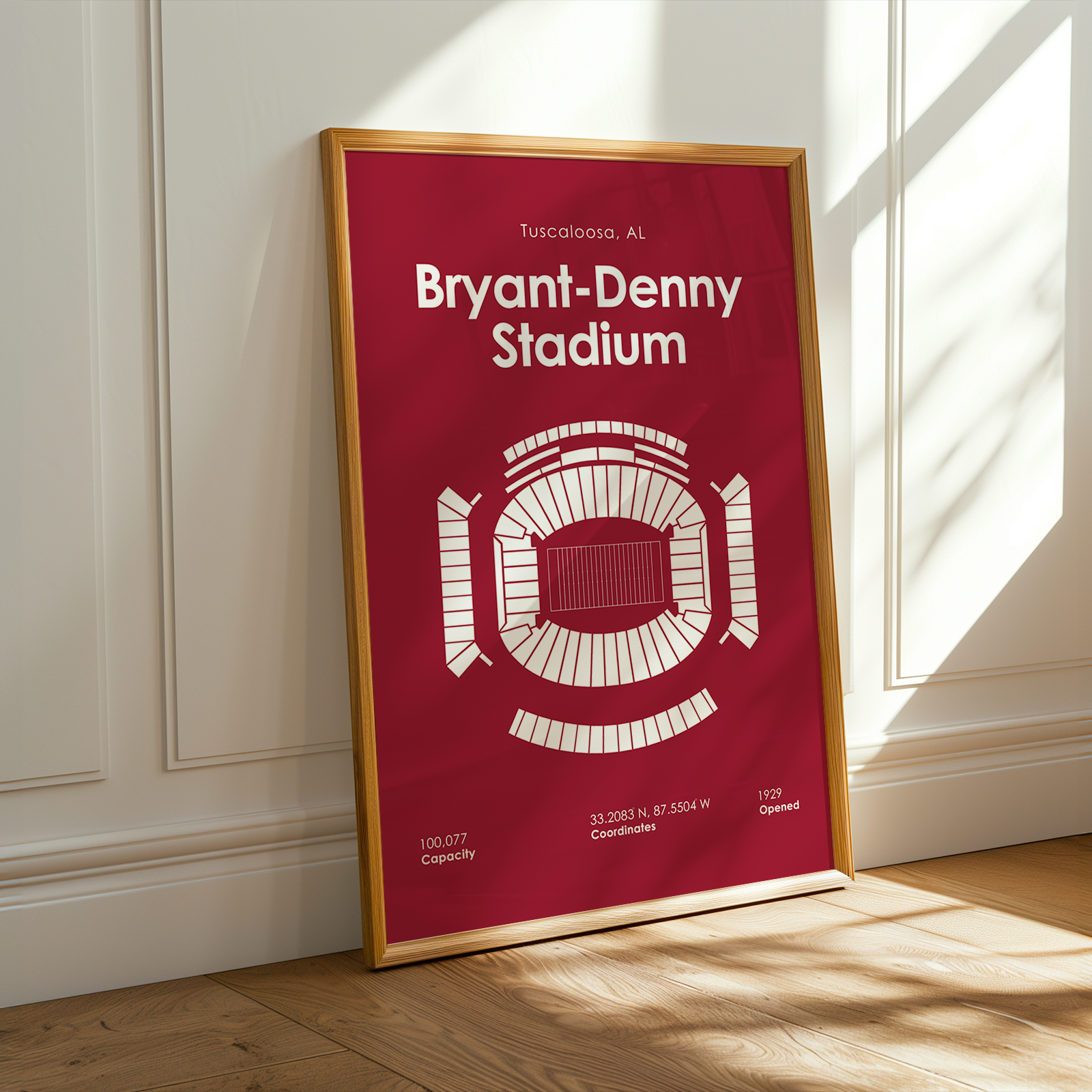 A framed sports poster of Bryant-Denny Stadium in Tuscon, Arizona, leaning against a white paneled wall on a wooden floor with warm sunlight streaming in from the side.