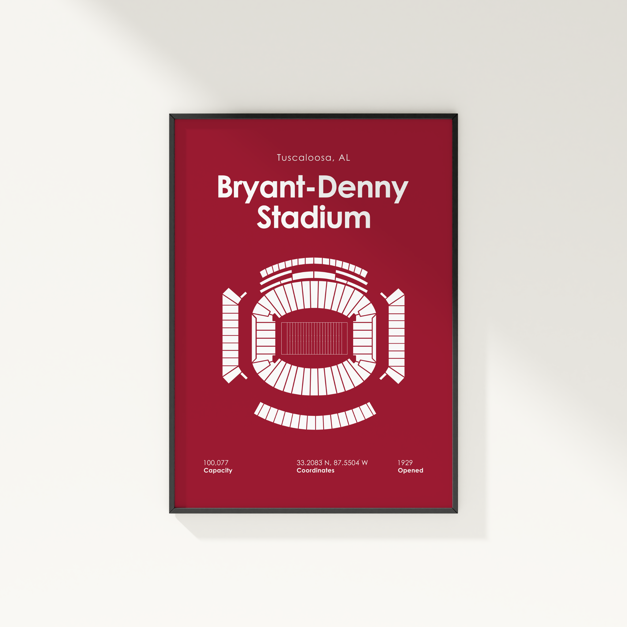 A framed vertical poster of Bryant-Denny Stadium in Tuscaloosa, AL, featuring a white stadium floor plan and key facts on a deep red background, shown hanging on a light beige wall.