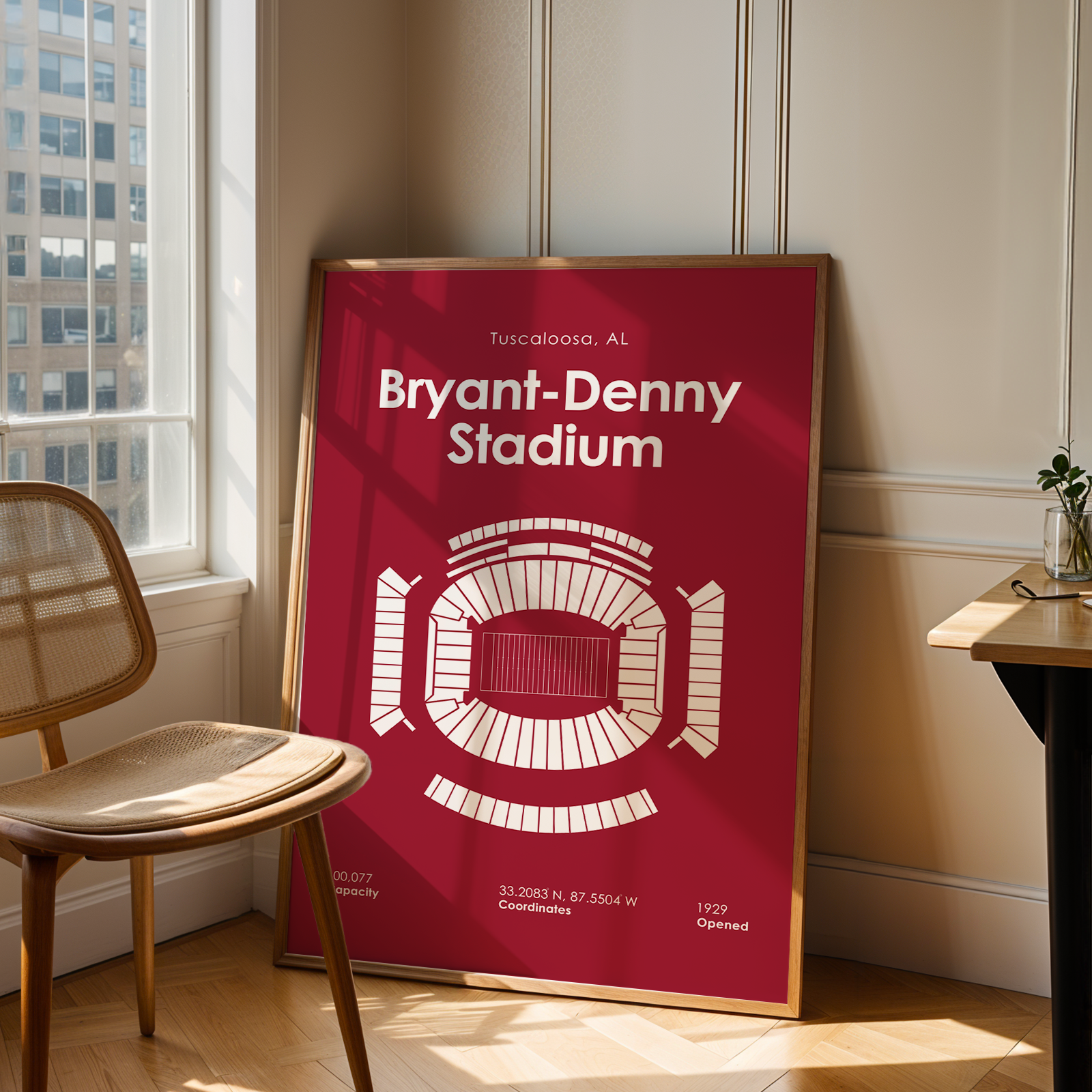 A framed art print of Bryant-Denny Stadium in Tuscaloosa, Alabama, leaning against a beige paneled wall in a sunlit room next to a wooden chair and desk.