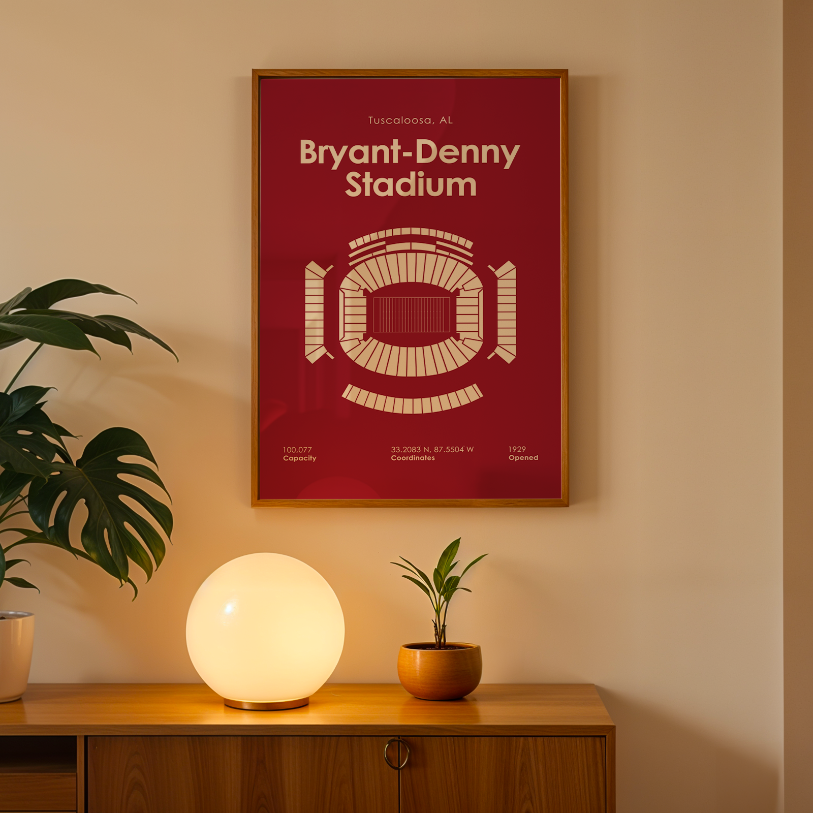 A framed poster of Bryant-Denny Stadium in Tuscon, Arizona, featuring a cream stadium layout on a deep red background, hanging on a beige wall above a wooden console table with a glowing globe lamp and potted plants.