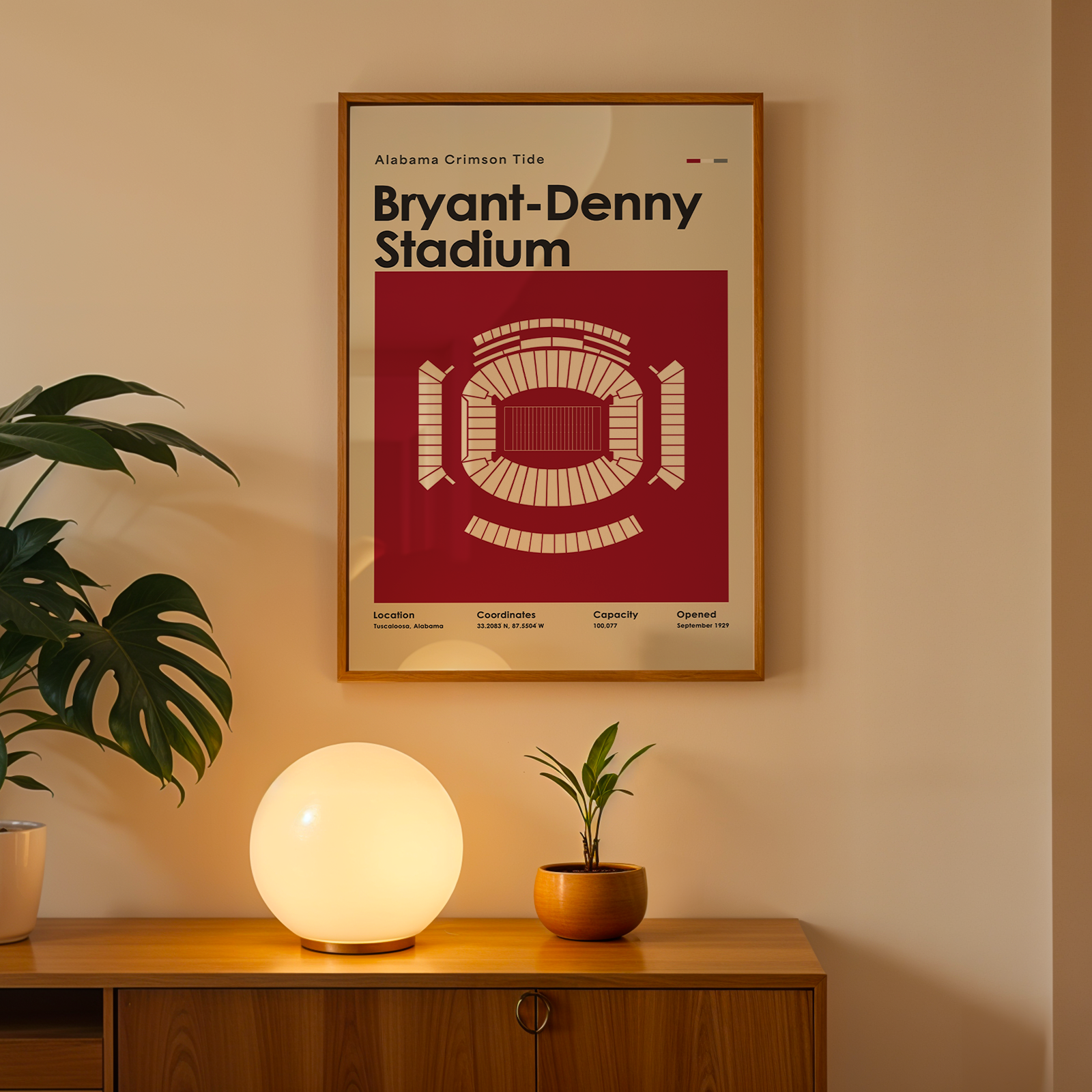 A framed Bryant-Denny Stadium poster hangs on a beige wall above a wooden console table. The poster features a red and cream stadium diagram with the text 'Bryant-Denny Stadium' and 'Alabama Crimson Tide.