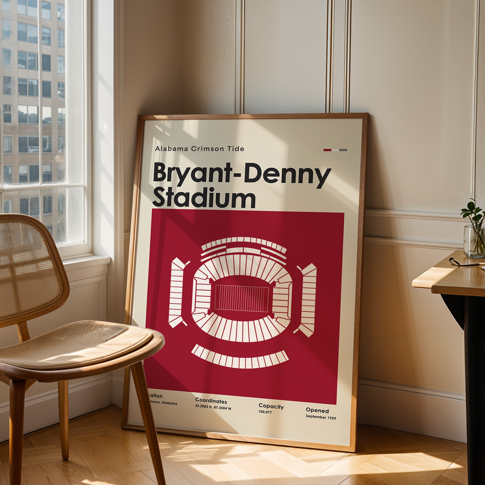 A framed poster of Bryant-Denny Stadium from the Alabama Crimson Tide leaning against a wall in a sunlit room next to a wooden chair and desk.