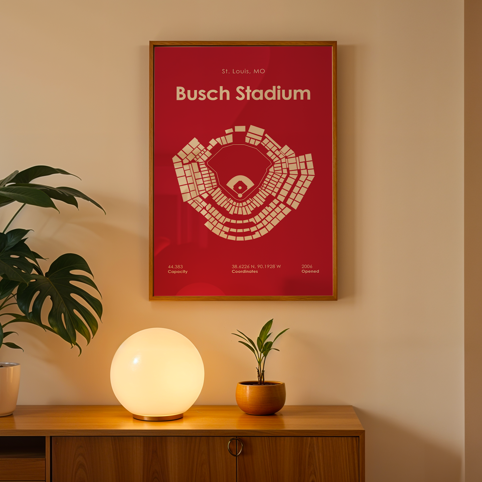Framed poster of Busch Stadium on a wall with a lamp and plant below.