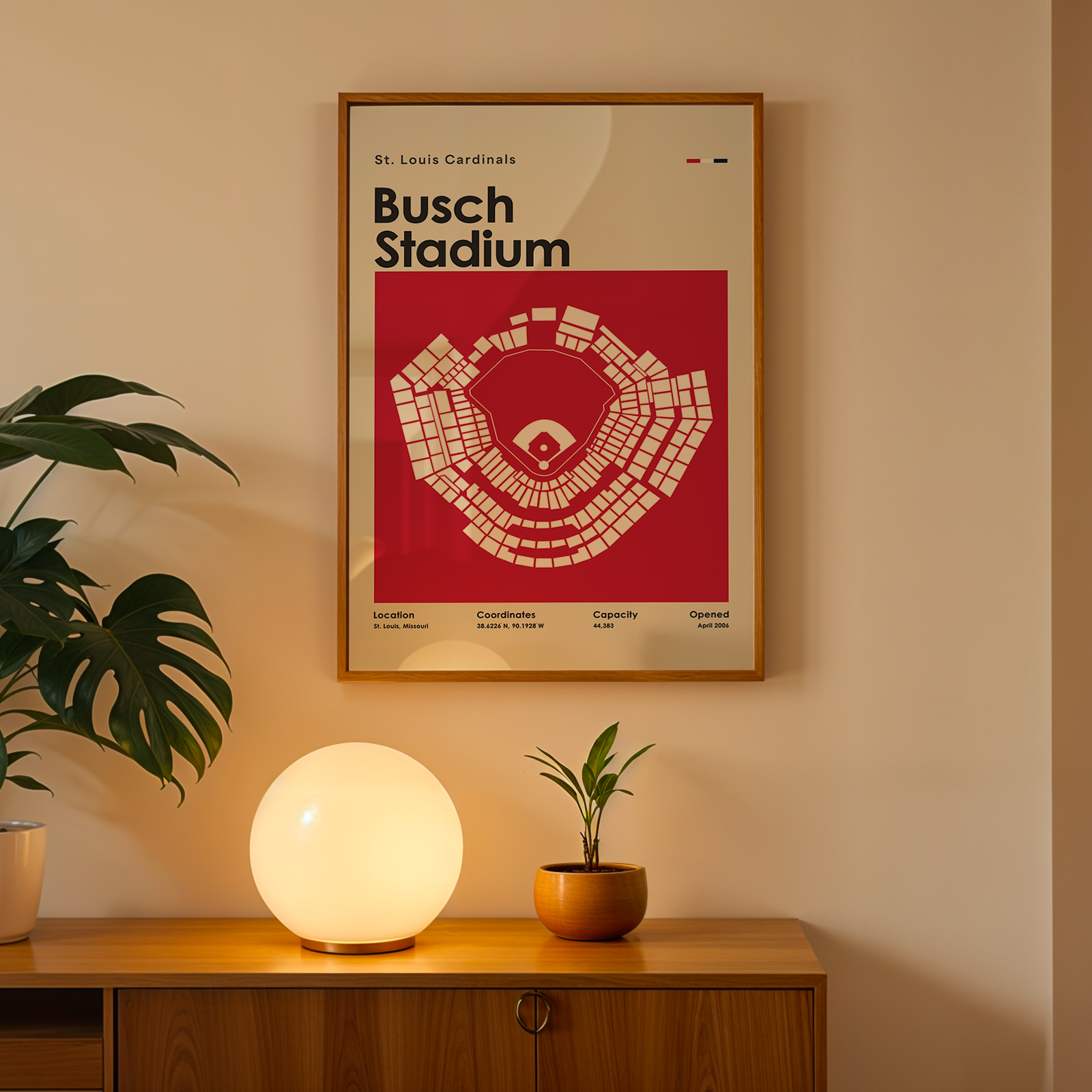 Framed poster of Busch Stadium above a wooden cabinet with decorative plants and a lamp.