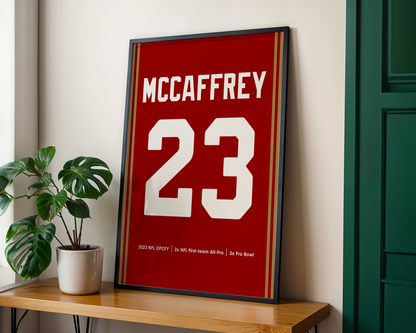 Framed poster with 'McCaffrey 23' on a wooden shelf with a plant and green door in the background