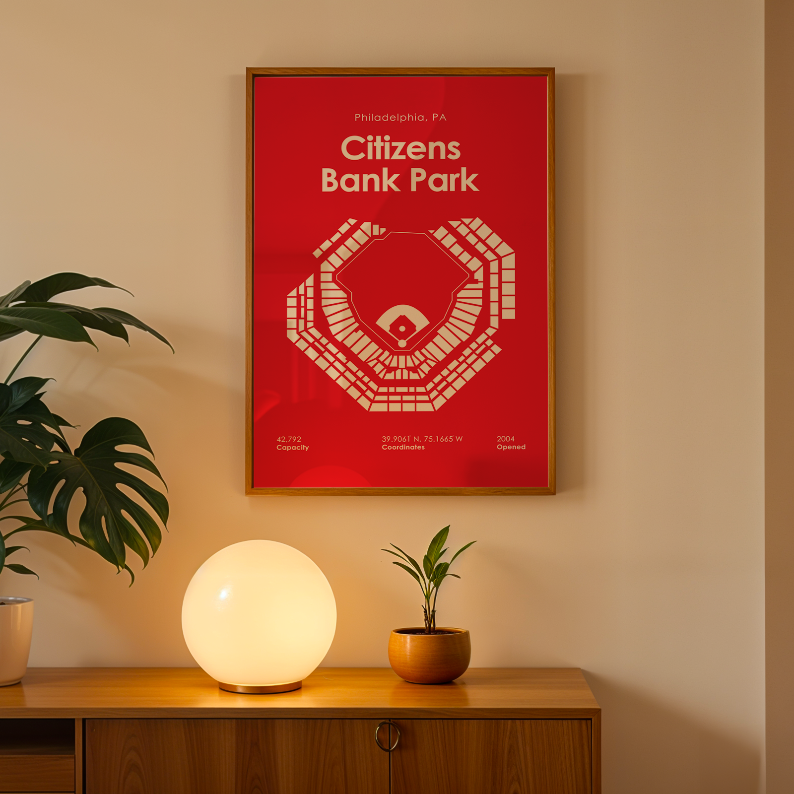 Framed poster of Citizens Bank Park on a wall with a plant and lamp below.