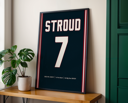 Framed sports jersey with 'STROUD' and number '7' on a wooden shelf.