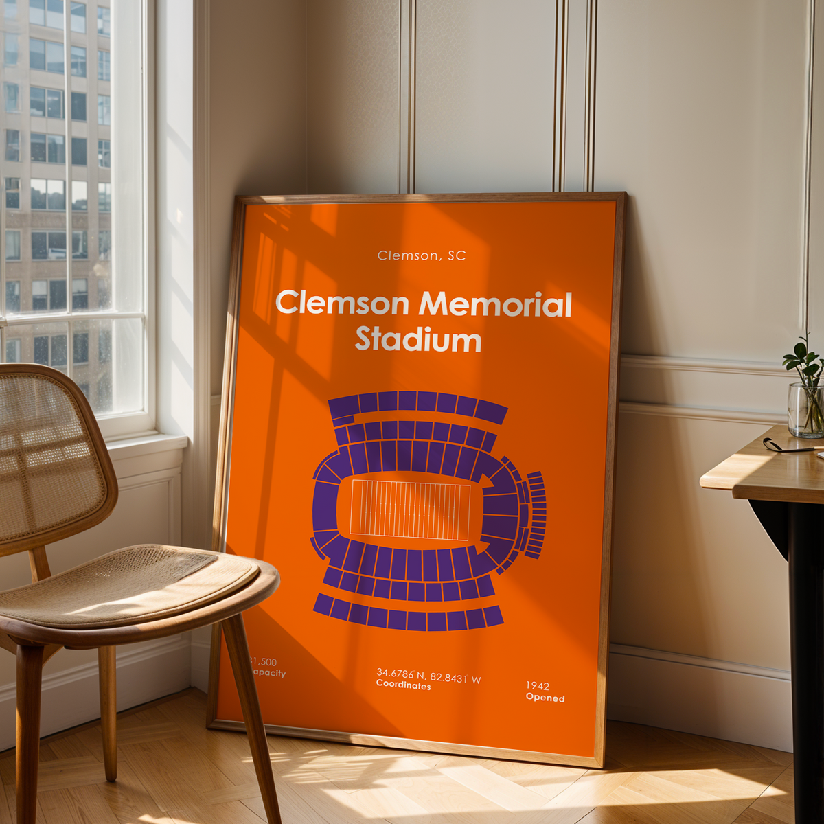 A framed Clemson University 'Clemson Memorial Stadium' art print leans against a wall in a sunlit room next to a wooden chair and a small desk.