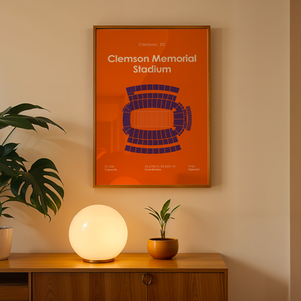 A framed Clemson Memorial Stadium poster with an orange background and dark blue stadium diagram, hanging on a beige wall above a wooden console table with a glowing globe lamp and potted plants.