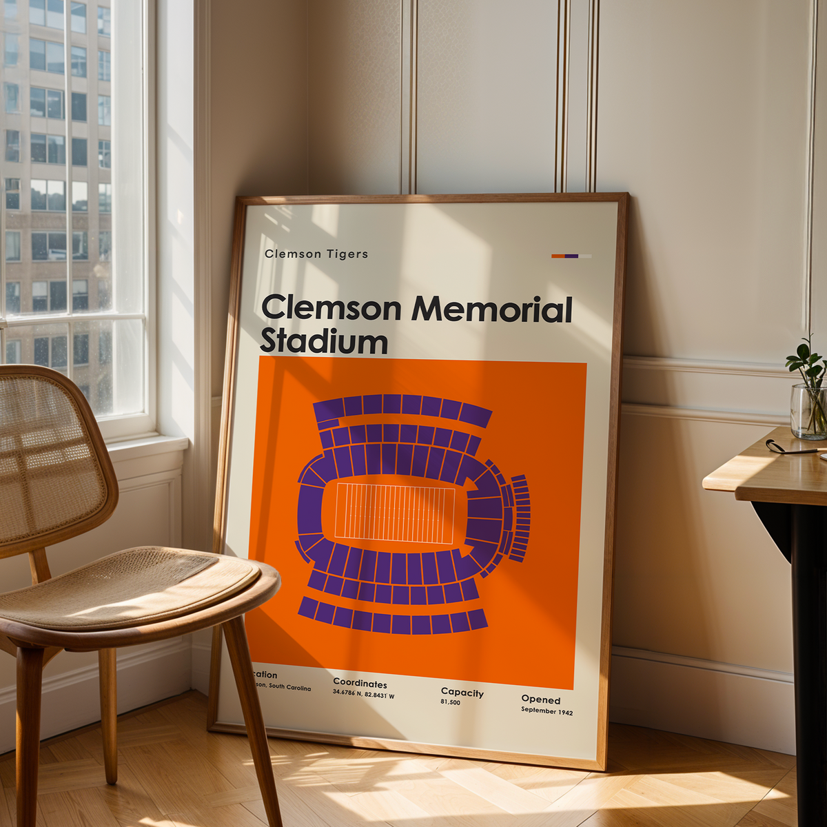 A framed Clemson Tigers stadium poster leaning against a wall in a sunlit room, next to a wooden chair and a small desk with a plant.