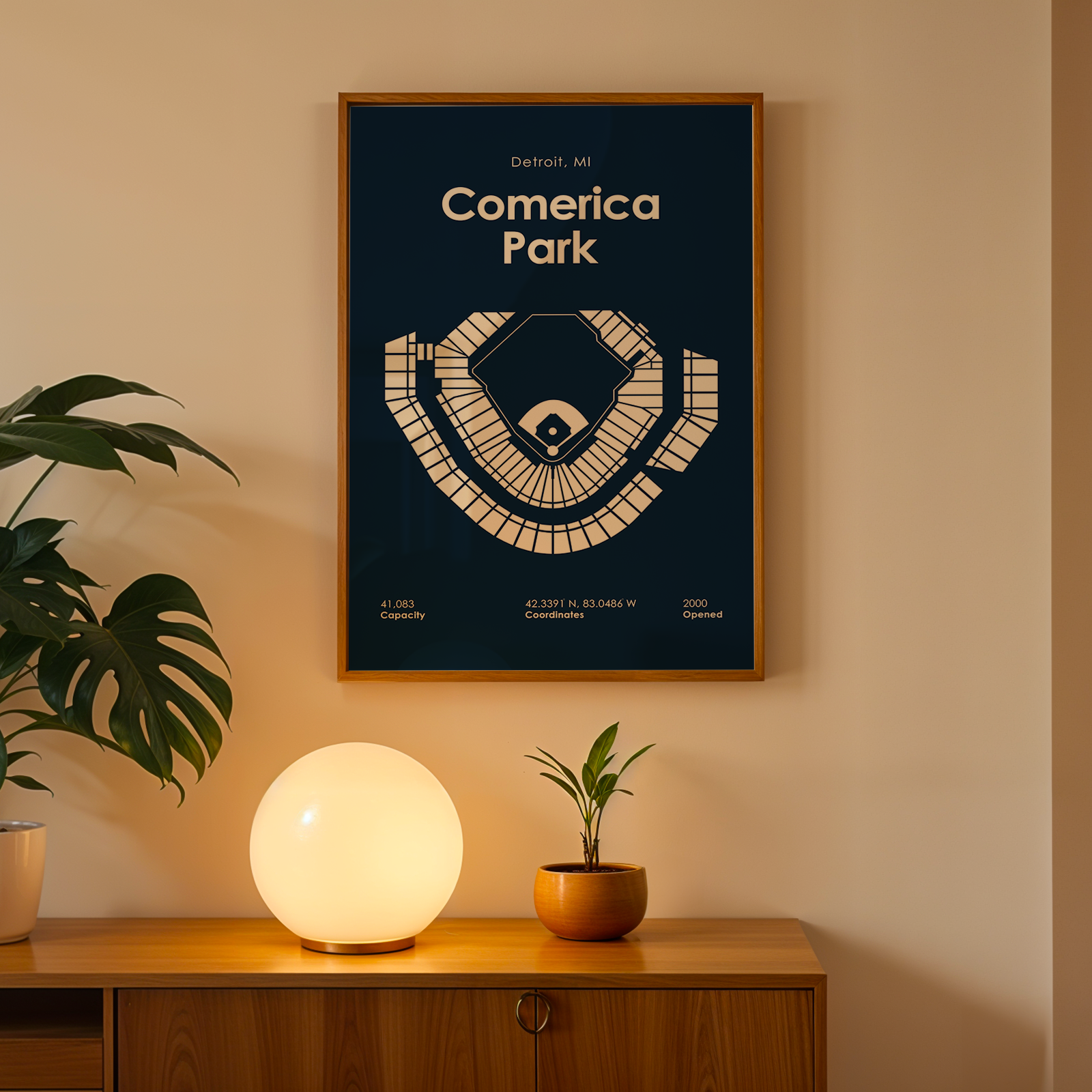 Framed poster of Comerica Park on a wall with a plant and lamp below.