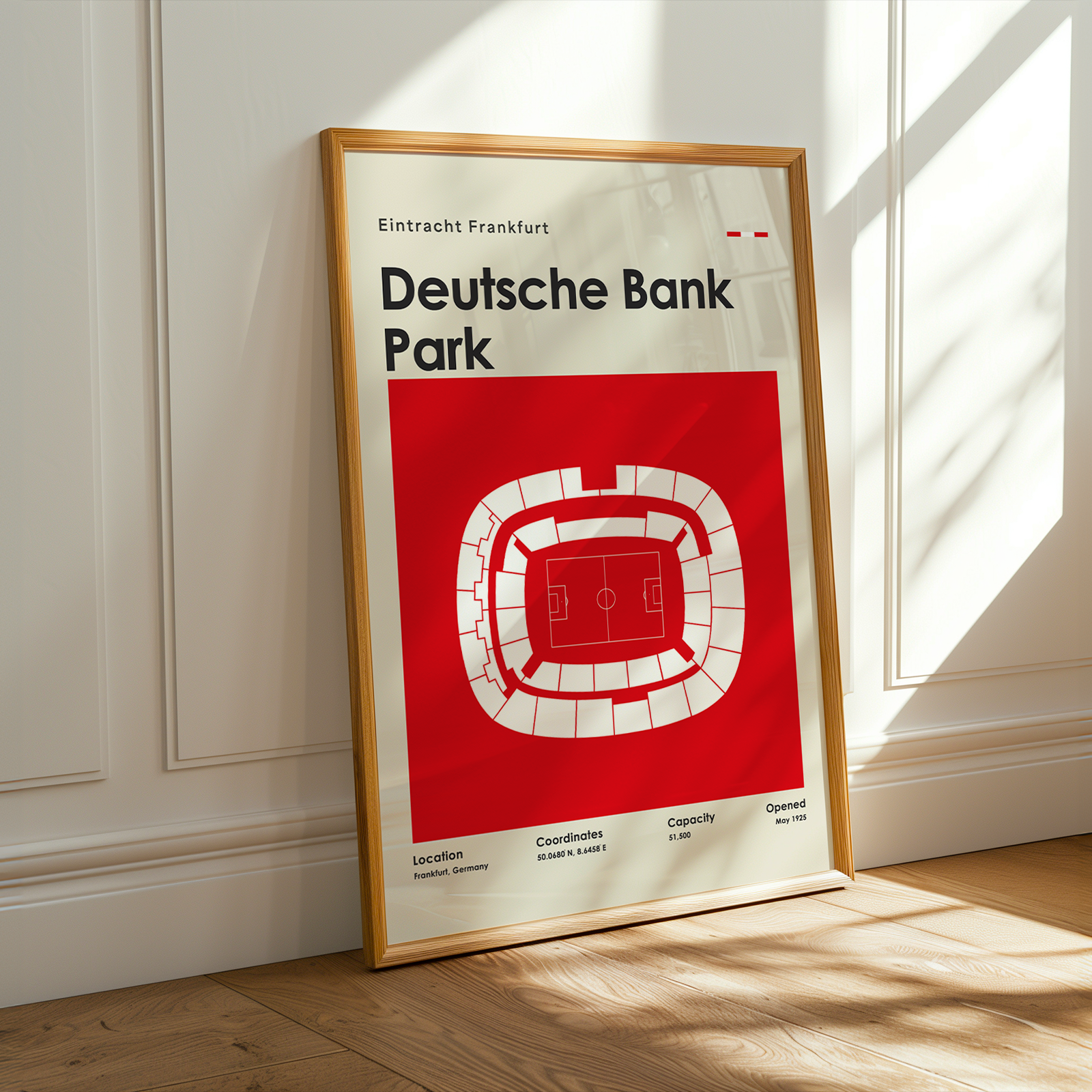 A framed poster of the Deutsche Bank Park stadium in Frankfurt, leaning against a white paneled wall on a wooden floor with warm sunlight streaming in.