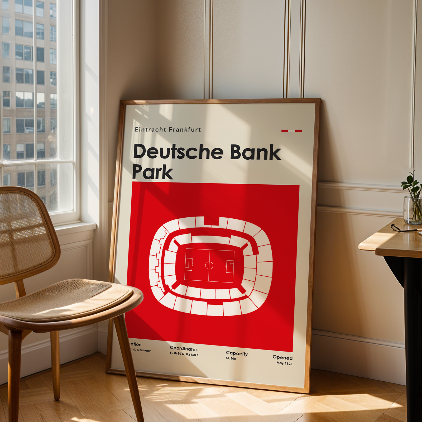 A framed poster of the Deutsche Bank Park stadium in Frankfurt, featuring a bold red and white graphic design, leaning against a beige paneled wall in a sunlit room next to a wooden chair and desk.
