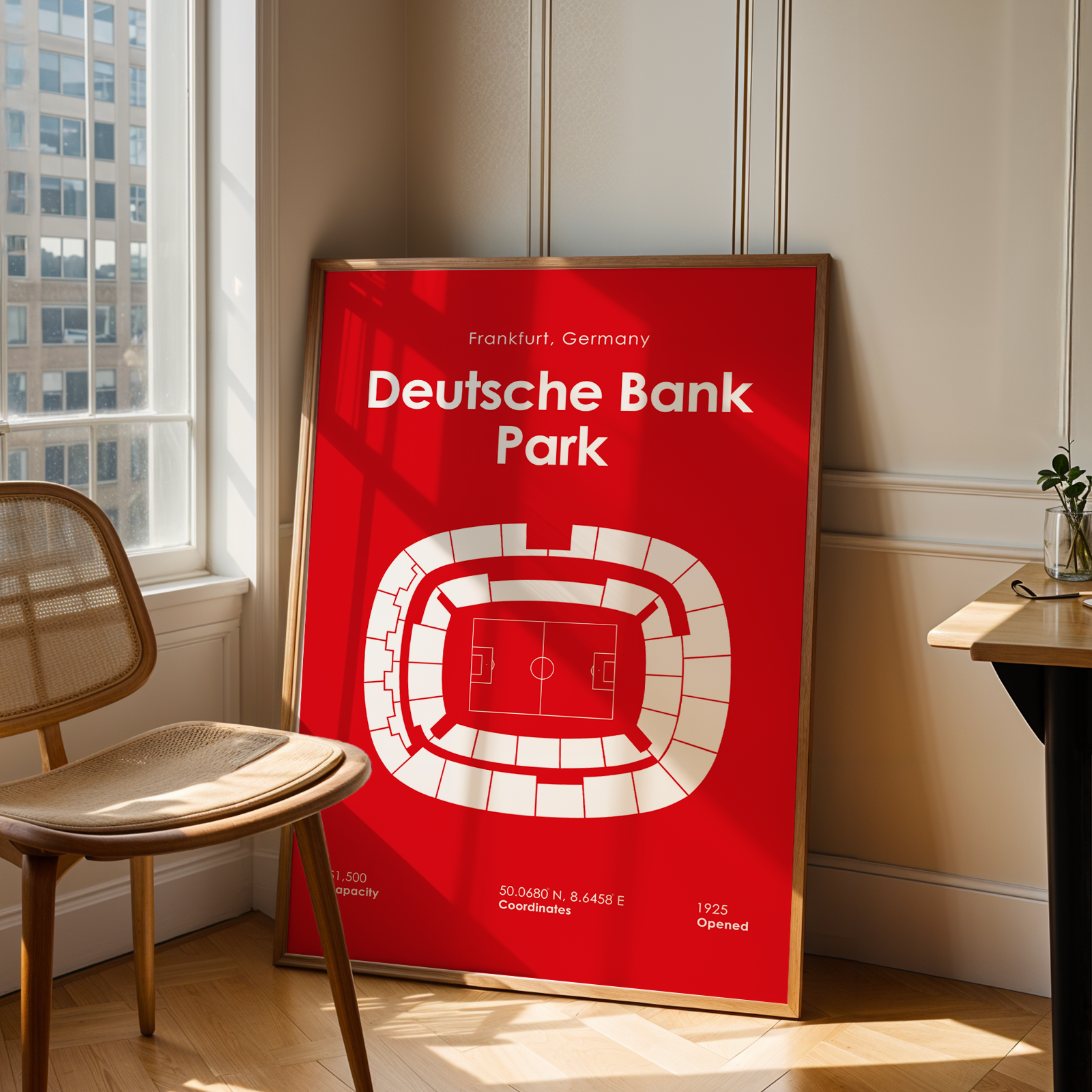 A framed red and white poster of Deutsche Bank Park in Frankfurt, Germany, leaning against a beige paneled wall in a modern room with a wooden chair and desk.