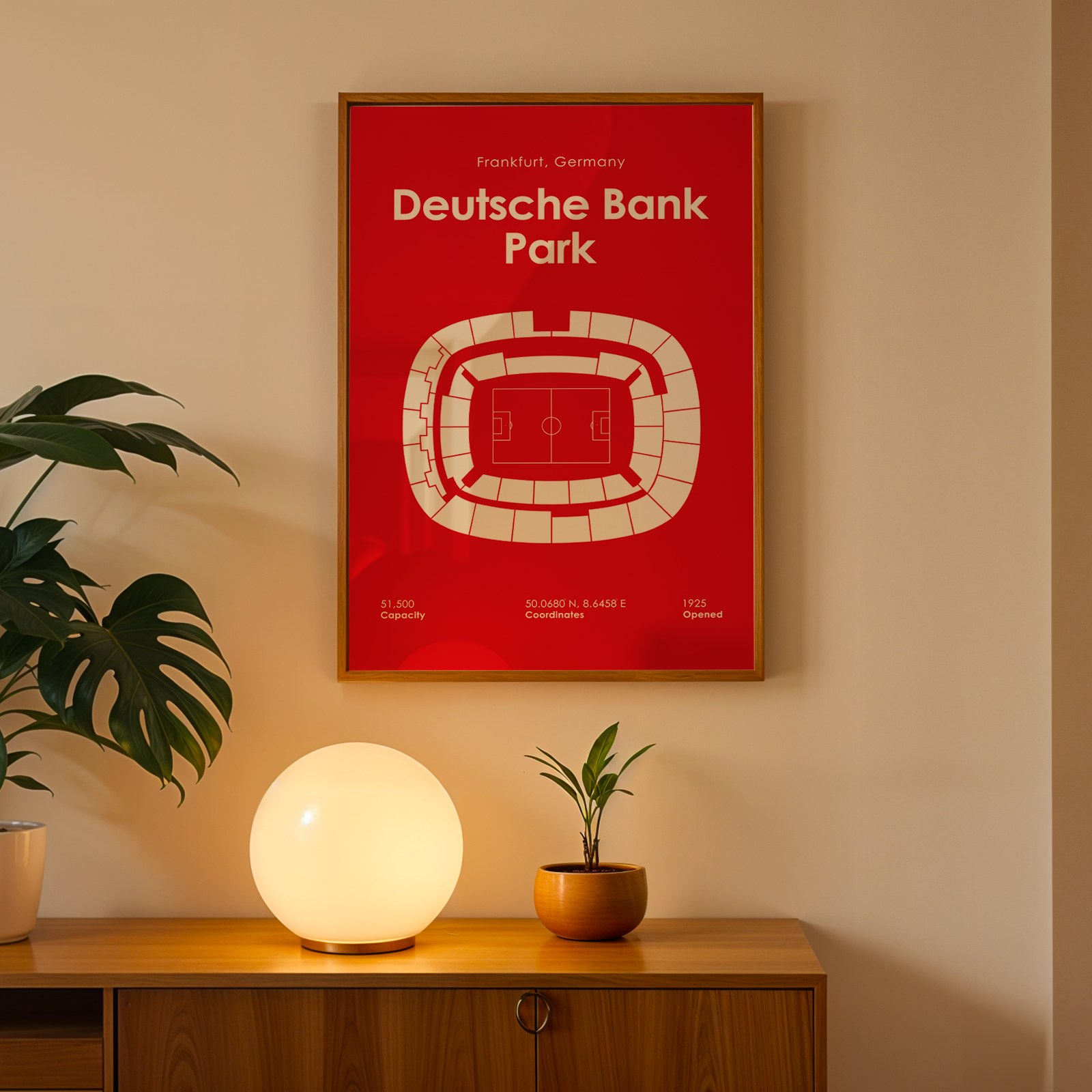 A framed red poster of Deutsche Bank Park football stadium in Frankfurt, Germany, hanging on a beige wall above a wooden console table with a glowing globe lamp and potted plants.