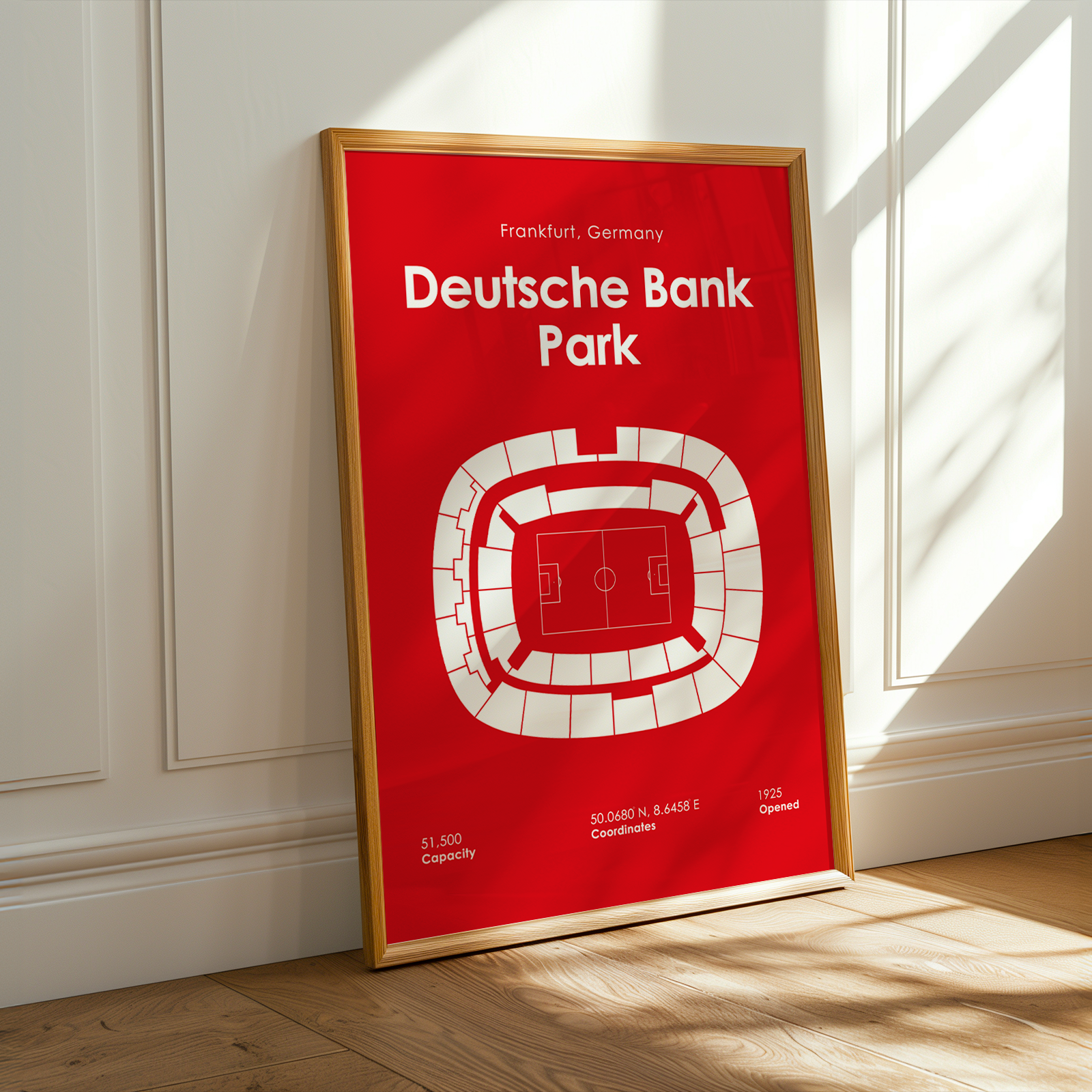 A framed red and white Deutsche Bank Park stadium map print leaning against a white paneled wall on a light wood floor, with warm sunlight casting shadows across the scene.